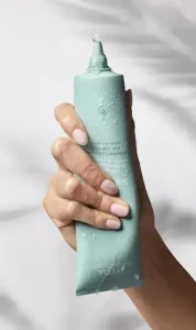 A hand holding a light green tube of botanical-hair-treatment with a small amount dispensed from the nozzle, against a soft, blurred background. | Simple Scissors Salon - Janesville, WI