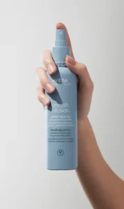 A hand holds a blue bottle of Aveda Smooth Infusion Perfect Blow Dry spray, showcasing this botanical-hair-treatment from aveda-hair-care against a plain light background. | Simple Scissors Salon - Janesville, WI
